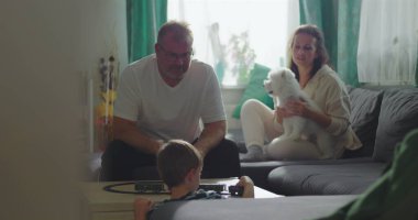 Baba kahve masasında oyuncak trenle oynayan çocuğu izliyor, anne arka planda küçük beyaz bir köpek tutuyor, aile bağı ve sakin bir oturma odasında oyun zamanı.