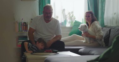 Baba ve çocuk sehpanın üzerinde oyuncak trenle etkileşime giriyor, anne arka planda beyaz köpeği tutuyor, yakın aile bağı ve kapalı alanda kaynaşma.