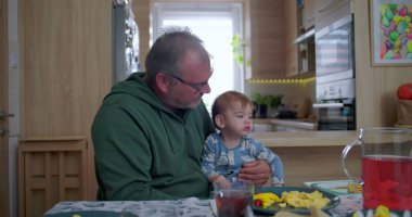 Baba aile yemeği sırasında bebeği kucağında tutuyor, bebek merakla yukarı bakıyor sıcak bir evde yemek masasında otururken.