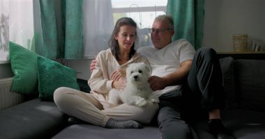 Küçük beyaz köpekle kanepede dinlenen, sakin ve samimi aile anlarının tadını çıkaran, doğal ışık ve yumuşak mobilyaları olan rahat oturma odası, sıcak atmosferi olan çift.