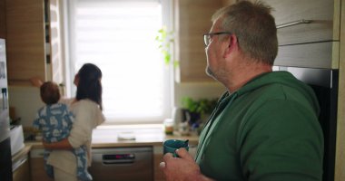 Anne bebeğini mutfakta tutarken bardaktan içen adam, aile sabahı rutini, ev yaşamında birden fazla işi olan ebeveynlerle sıcak aile sahnesi.