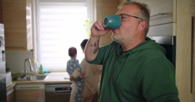 Annesi buzdolabını kollarında bebekle kontrol ederken elinde bardak tutan bir adam, tipik bir sabah rutini sırasında sıradan bir aile anı, sıcak ve besleyici ebeveynlik sahnesi.