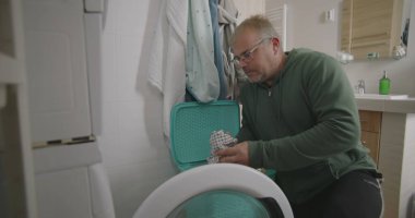 Çamaşır makinesinin önünde çömelmiş bir adam, çamaşırları küçük bir banyodaki çamaşır makinesine yüklüyor, ev işlerini odaklanarak ve verimlilikle yapıyor.