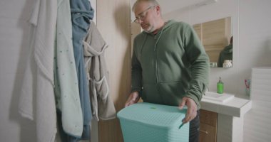 Çamaşır makinesinin önünde çömelmiş bir adam, çamaşırları küçük bir banyodaki çamaşır makinesine yüklüyor, ev işlerini odaklanarak ve verimlilikle yapıyor.