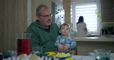 Babam yemek masasında renkli yemek ve içeceklerle bebeği tutarken annem de mutfakta, sıcak aile ortamında sıcak aile ortamını hazırlıyor.