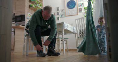 Babam, sıcak ahşap döşemeleri ve doğal ışıklandırması olan sıcak bir aile yemek odasında, emzik ile perdelerin yanında duran bir çocuğa bakarken botlarını bağlıyor.