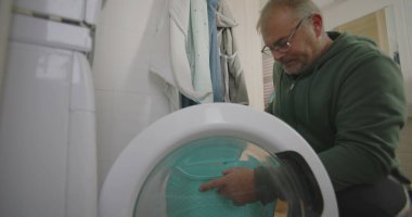 Çamaşır makinesinin önünde diz çökmüş bir adam, temiz havluları ve parlak döşeli duvarları olan modern bir banyoda çamaşırları dikkatlice yüklüyor.