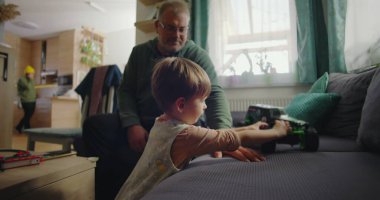Babası gülümserken oğlu kanepede yeşil bir oyuncak kamyonla oynuyor. Sıcak ve rahat bir oturma odasında, ahşap mobilyaları ve canlı bir iç mekanı var.