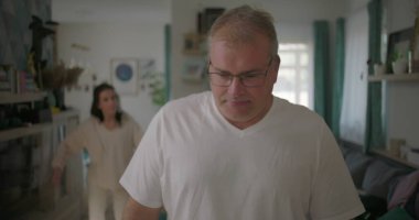 Oturma odasında hararetli tartışmalar yaşayan bir çift, görünürde üzgün duran bir adam, arka planda duygusal hareketler yapan bir kadın, ilişki çatışması sırasında gergin bir an.