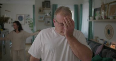 Oturma odasında hararetli tartışmalar yaşayan bir çift, görünürde üzgün duran bir adam, arka planda duygusal hareketler yapan bir kadın, ilişki çatışması sırasında gergin bir an.