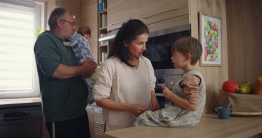 Modern mutfaktaki aile etkileşimi, baba bebeğini kucağında tutarken annesinin tezgahta oturması, sıcak aile bağlarının kurulması.