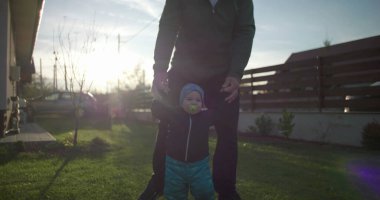 Baba, çocuğun aile evinin yakınındaki çimenlikte yürümesine yardım ediyor. Çocuğun hareketini ve dengesini destekliyor. Bağlanmayı ve sevgiyi teşvik ediyor.