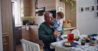 Baba kucağında bebek ve gülümsüyor. Sıcak bir mutfak ortamında neşeli ve sevecen bir an yaşıyor.