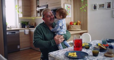 Baba kucağında bebek ve gülümsüyor. Sıcak bir mutfak ortamında neşeli ve sevecen bir an yaşıyor.