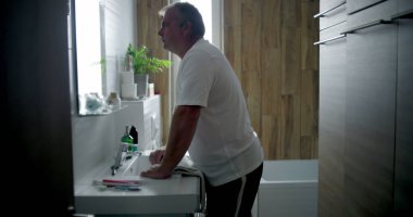 Profil görünümlü bir adam banyoda öne doğru eğiliyor, derin düşüncelere dalmış, içi beyaz fayanslarla dolu bir banyo ve ahşap kaplama, sessiz ve yansıtıcı kişisel bir an.