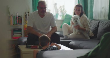 Baba kahve masasında oyuncak trenle oynayan çocuğu izliyor, anne arka planda küçük beyaz bir köpek tutuyor, aile bağı ve sakin bir oturma odasında oyun zamanı.