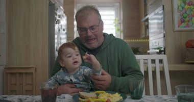 Babam yemek masasında kucağında bebeğini tutarken gülümsüyor, sıcak ve sevgi dolu bir aile anı yaratıyor.