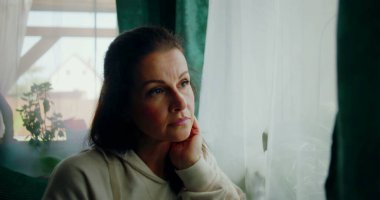 Sakin ve düşünceli bir ifadeyle dışarı bakan bir kadının yakın çekimi, çenesini elinde dinlendirirken, yumuşak perdelerle çerçevelenmiş ve rahat bir iç mekan.