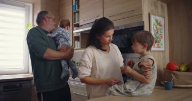 Modern mutfaktaki aile etkileşimi, baba bebeğini kucağında tutarken annesinin tezgahta oturması, sıcak aile bağlarının kurulması.