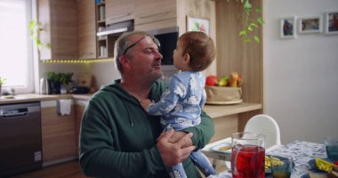 Baba kucağında bebek ve gülümsüyor. Sıcak bir mutfak ortamında neşeli ve sevecen bir an yaşıyor.