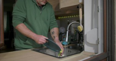 Bir adam mutfak lavabosundaki musluk suyunun altında mavi bir tabağı duruluyor, günlük ev işleri sırasında bulaşık takımlarını temizlemeye odaklanıyor.