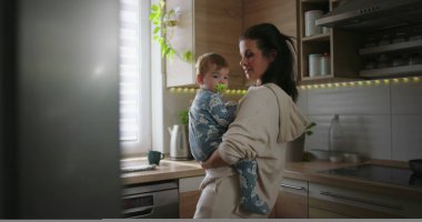 Anne mutfak tezgahının yanında bebeği tutuyor, çocuğa gülümsüyor ve bağ kuruyor, günlük aile rutini sırasında sıcak ve şefkatli ebeveyn anlarını yaşıyor..