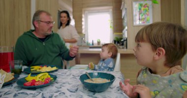 Aile kahvaltısı sırasında masada gülümseyen küçük bir çocuk, sandalyesinde yemek tutan küçük bir çocuk, karnını doyuran bir baba, arka planda duran bir anne.