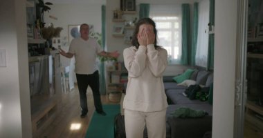 Kadın, arka planda bir erkekle tartışırken kararlı bir şekilde oturma odasında duruyor. Öfkeyle el kol hareketi yapıyor, bir çiftin ilişkilerindeki gerginliği ve çatışmayı yakalıyor.