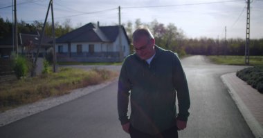 Orta yaşlı bir adam, sakin bir sokakta elleri ceplerinde, sakin bir duruş ve sakin bir mahalle atmosferi sergiliyor.