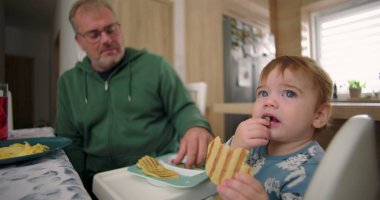 Bebek kahvaltıda yüksek sandalyede otururken waffle yiyor, baba sıcak bir ilişki kuruyor ve sıcak bir ev mutfağı kuruyor.