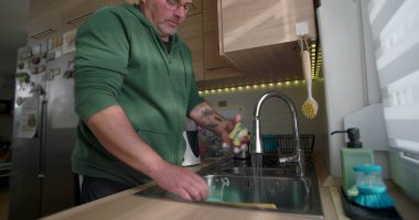 Bir adam mutfak lavabosundaki musluk suyunun altında mavi bir tabağı duruluyor, günlük ev işleri sırasında bulaşık takımlarını temizlemeye odaklanıyor.