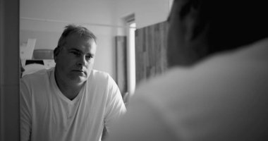 Banyo aynasında yansıyan adam, siyah beyaz ton, düşünceli ifade, eller tezgahta, ahşap banyo detayları arka planda, sakin ve sakin atmosfer