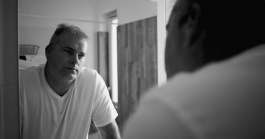 Banyo aynasında yansıyan adam, siyah beyaz ton, düşünceli ifade, eller tezgahta, ahşap banyo detayları arka planda, sakin ve sakin atmosfer