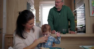 Anne, bebeğin yemek masasında otururken su içmesine yardım ediyor, baba arka planda dikiliyor, sıcak bir aile mutfağında.