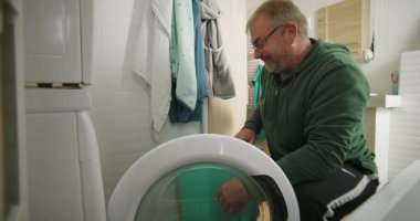 Çamaşır makinesine çamaşır sepeti ve temiz ve modern banyo ortamında havluları koymaya hazırlanırken gülümseyen bir adam.
