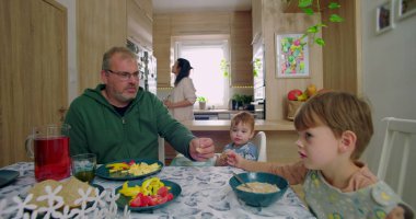 Aile kahvaltısı sırasında masada gülümseyen küçük bir çocuk, sandalyesinde yemek tutan küçük bir çocuk, karnını doyuran bir baba, arka planda duran bir anne.