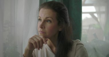 Pencerenin yanındaki rahat bir koltukta oturan kadın, kolunu yastığa koyuyor, sakin bir oturma odasında düşünceli bir ifadeyle dışarı bakıyor.