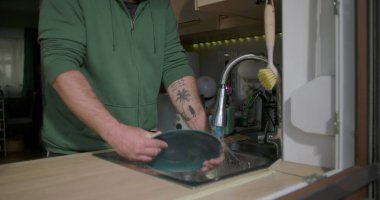 Bir adam mutfak lavabosundaki musluk suyunun altında mavi bir tabağı duruluyor, günlük ev işleri sırasında bulaşık takımlarını temizlemeye odaklanıyor.