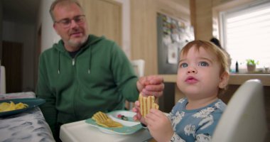 Bebek kahvaltıda yüksek sandalyede otururken waffle yiyor, baba sıcak bir ilişki kuruyor ve sıcak bir ev mutfağı kuruyor.