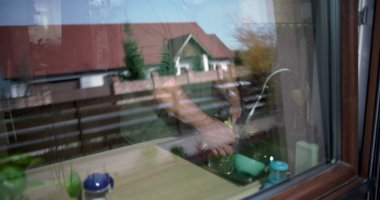 Yansıtıcı bir pencereden bulaşıkları yıkayan, iç ve dış perspektiflerle harmanlayan, günlük ev aktivitesi, doğal çevreye sahip modern ev