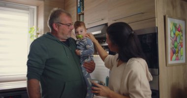 Anne, modern mutfakta babayla konuşurken bebeğin kafasına şefkatle dokunuyor. Günlük ev ortamında sıcak aile etkileşimi..