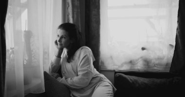 Kanepede oturan bir kadın, düşünceli bir şekilde pencereden dışarı bakıyor, etrafını bitkiler ve perdeler sarmış, tek renkli tonlarda, huzurlu ev ortamını sessizce yansıtıyor.