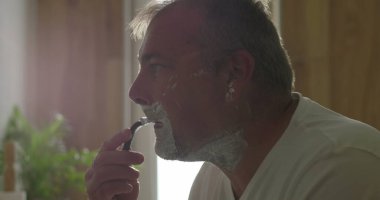 Jiletle tıraş olan, başını hafifçe eğen, temiz bir tıraş için baskı uygulayan, yüzünde köpük olan, arka planda banyo detayları, hijyen ve bakım yapan bir adam.