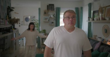 Erkek, arka plandaki kadınla tartışırken gözle görülür şekilde üzgün, oturma odasında ilişki gerilimi, duygusal ve dramatik anlaşmazlık anı