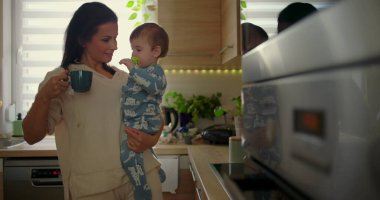 Anne mutfakta bebeği tutarken çocuk sıcak fincana uzanır, günlük aile rutini sırasında koruyucu ebeveynlik anı, sıcak bir evde bakım ve güvenlik.