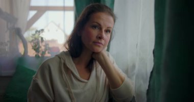 Çenesi elinde dinlenen bir kadın, derin bir düşünceyle pencereden dışarı bakıyor, yumuşak perdelerle çevrili ve evdeki sıcak elementlerle çevrili.