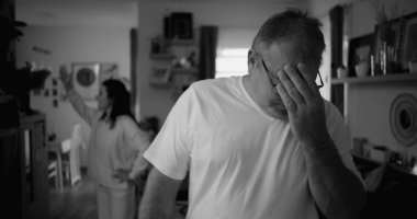 Tartışma sırasında yoğun ifadeli erkek portresi, oturma odasında arka planda kadın, duygusal gerilim ve gergin ilişki dinamiklerine odaklan, siyah-beyaz