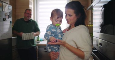 Anne kucağında bebek, baba arka planda çalışırken aile dinamiklerini ve günlük rutinlerini gözler önüne seriyor.