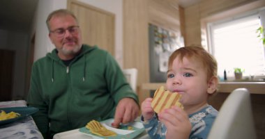 Bebek kahvaltıda yüksek sandalyede otururken waffle yiyor, baba sıcak bir ilişki kuruyor ve sıcak bir ev mutfağı kuruyor.
