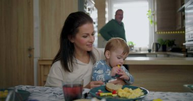 Anne gülümserken, bebek kahvaltıda sarı dolmalık biber ararken sıcak ve besleyici atmosferli sıcak bir mutfakta, aile yemeği eşliğinde kaynaşıyor.